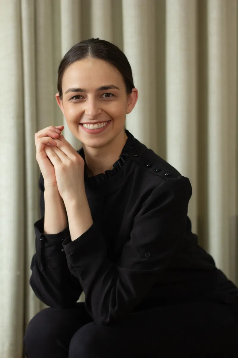 Retrato de Lara Turatti Issa, sorrindo, de terno branco e óculos, diante de uma parede clara